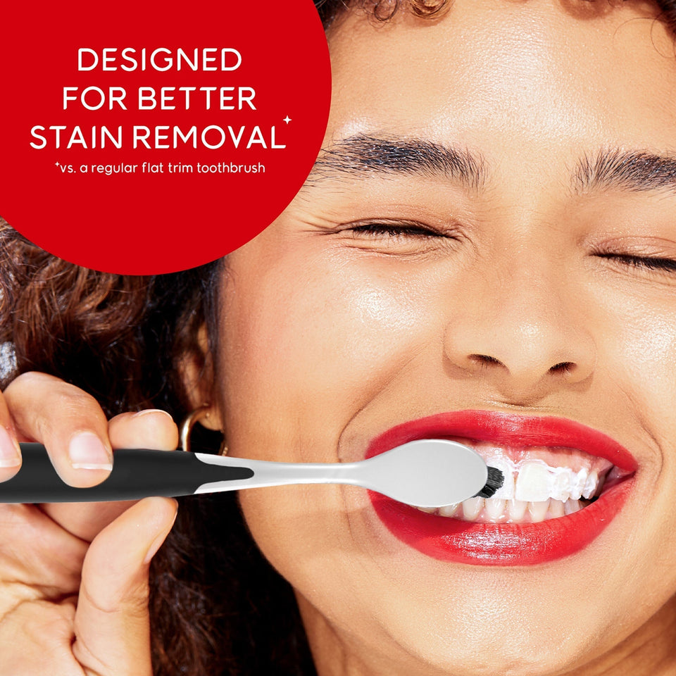 A woman smiling and brushing her teeth with an Optic White Pro Series Charcoal Manual Toothbrush. In text: Designed for better stain removal*. *Vs. a regular flat trim toothbrush.