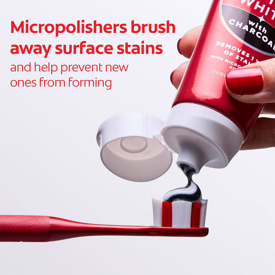 A hand squeezing Colgate Optic White with Charcoal Teeth Whitening Toothpaste onto a red toothbrush. In text: Micropolishers brush away surface stains and help prevent new ones from forming. 