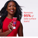 A woman with white teeth smiling and holding a tube of Colgate Optic White Purple Toothpaste for Teeth Whitening. In text: Removes 95% of daily surface stains*. *After 2 weeks of twice daily use. 
